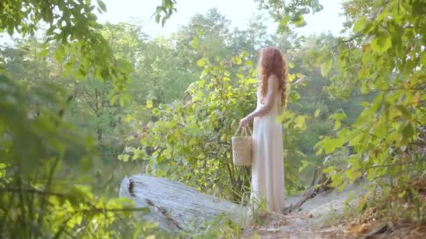 Beautiful slim caucasian woman with red hair standing on the riverbank holding basket and touching her long hair. Leisure outdoors. Connection with nature. Rural life — Stock Video