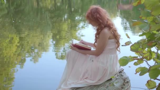 Рыжая привлекательная кавказская девушка читает книгу в темно-красной обложке на берегу озера в летнем лесу. Сказочная женщина, увлеченная поэзией . — стоковое видео