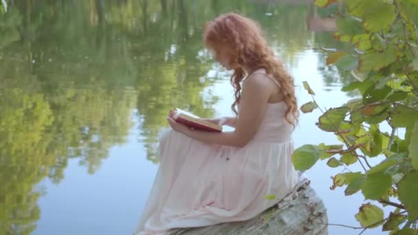 Seitenansicht des rothaarigen attraktiven kaukasischen Mädchens, das das Buch in dunkelrotem Einband am Ufer des Sees im Sommerwald liest. Fee mit Lust auf Poesie.