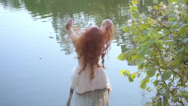 Beyaz bir kadının uzun kızıl saçlarını okşayıp sonbahar parkında gölün kıyısında güneşin tadını çıkarıyor.. — Stok video