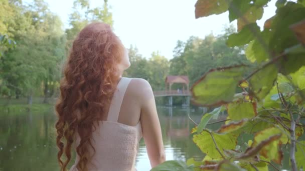 Vista trasera de una mujer caucásica increíblemente encantadora tomando el sol en la orilla del lago en el parque de otoño. Chica pelirroja atractiva girando a la cámara y sonriendo suavemente . — Vídeos de Stock