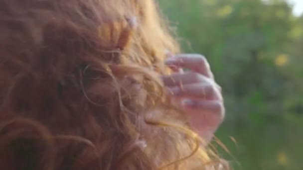 Close-up de jovem mulher caucasiana acariciando seu longo cabelo vermelho e desfrutando do sol na margem do lago no parque de outono — Vídeo de Stock