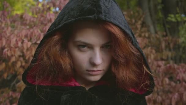 Faccia da primo piano di attraente donna caucasica dai capelli rossi con un abito nero che guarda la telecamera. Strega cattiva nella foresta autunnale. Fiaba, misticismo. Halloween — Video Stock