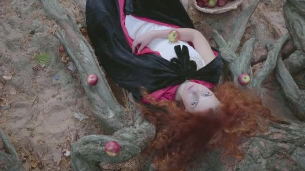 Atractiva mujer caucásica pelirroja en vestido blanco y vestido negro tendida en el árbol rodeado de manzanas rojas, mirando a la cámara. Bastante joven bruja descansando en el bosque de otoño . — Vídeo de stock