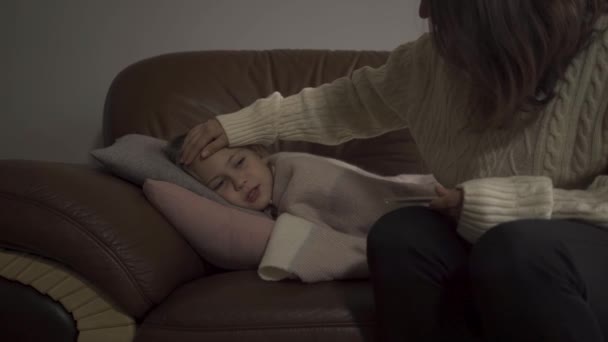 Молодая кавказская мать сидит дома со своей больной дочерью. Маленькая хорошенькая девочка лежит под одеялом с лихорадкой. Концепция здоровья, болезни, болезни, простуды, лечения . — стоковое видео