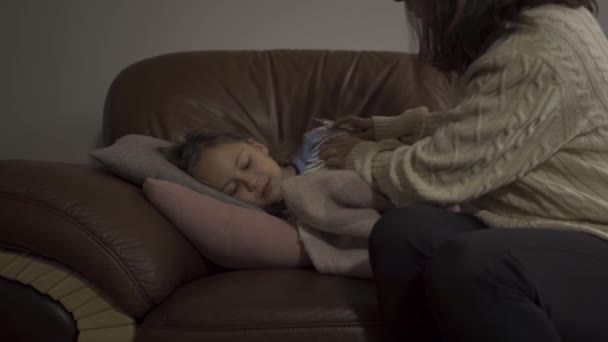 Jovem mãe caucasiana em suéter branco levando a temperatura de sua filha doente em casa. Menina bonita pequena deitada sob o cobertor quente com febre . — Vídeo de Stock