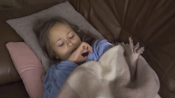 年轻的白种女孩穿着蓝色毛衣躺在温暖的毯子下，摄取温度。小病黑发孩子的特写. — 图库视频影像