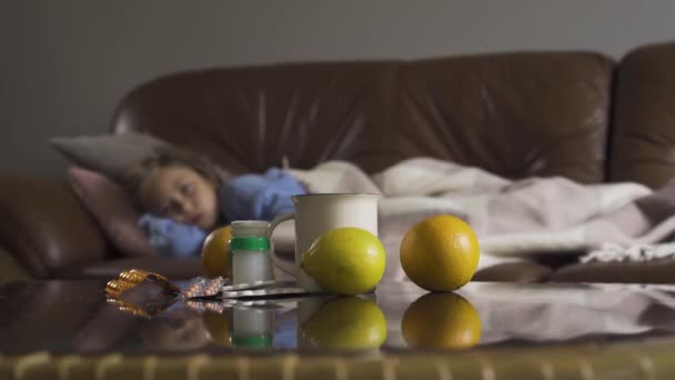 Kranke junge kaukasische Mädchen hustet, während sie zu Hause unter einer Decke im Hintergrund liegt. Tabletten, Zitronen und Teetasse auf dem Tisch im Vordergrund. Gesundheitsbegriff, Krankheit, Krankheit, Erkältung, Behandlung — Stockvideo