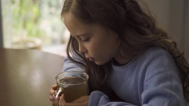 Маленькая белая девочка с длинными вьющимися волосами брюнетки, сидящая за чашкой ромашкового чая. Милый грустный ребенок, одетый в синий свитер, ждет своего горячего напитка, чтобы остыть . — стоковое видео