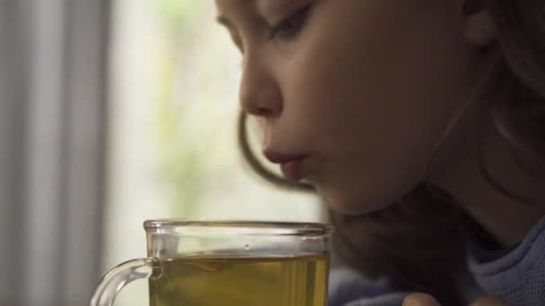 Close-up gezicht van klein meisje waait op de kruidenthee voordat het drinken terwijl zittend aan de tafel thuis. Het kind genieten van thee voor verkoudheid in de keuken. Zorgeloze kindertijd. Concept van de gezondheidszorg — Stockvideo