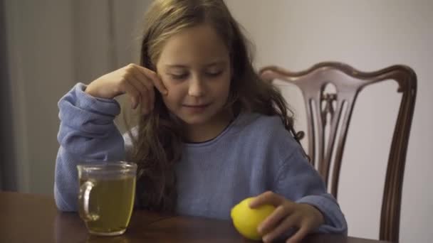 Närbild av liten flicka som håller citron medan du dricker örtte. Barn som sitter vid bordet hemma. Kid njuter av te för förkylningar i köket. Bekymmerslös barndom. Begreppet hälso-och sjukvård — Stockvideo