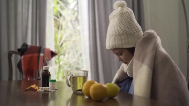 Schattige kleine Kaukasische meisje in warme witte muts en sjaal die zich met een deken bedekt. Kind zittend aan tafel met pillen, citroen en sinaasappelen. Begrip ziekte, ziekte, behandeling. — Stockvideo