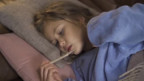 Young caucasian girl in blue sweater taking temperature. Close-up of a little sick brunette child laying under the warm blanket at home. — Stock Video