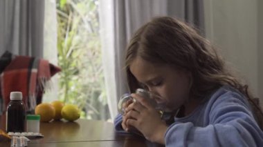 Evde masada oturan papatya çayı içen sevimli küçük bir kızın yakın çekim. Haplar, limon ve portakal, çocuğun yanındaki masada yatıyor. Sağlık kavramı.