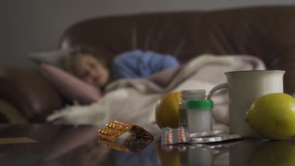 Маленькая грустная девочка лежит дома под одеялом и кашляет на заднем плане. Таблетки, лимоны и чашку чая на столе на переднем плане. Здоровье, болезнь, болезнь, простуда, лечение — стоковое видео