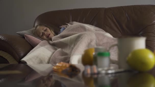 Tos niña tendida en el fondo con una mesa con pastillas, limones y una taza de té en primer plano. Niño caucásico en tratamiento en casa. Centrado en el fondo . — Vídeo de stock