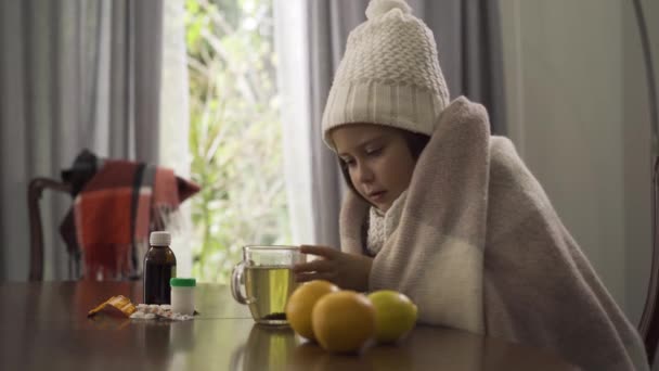 Close-up van Little Girl in winter muts en sjaal bedekt met deken drinken thee terwijl zittend aan de tafel thuis. Pillen, citroen en sinaasappelen zijn op de tafel. Begrip ziekte, ziekte, behandeling — Stockvideo