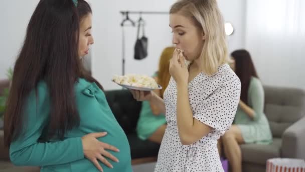 Twee aantrekkelijke Kaukasische zwangere vrouwen eten snoep en strelen hun buiken. Aanstaande vrouw en haar vriend hebben samen plezier. Jonge Kaukasische meisjes het hebben van pre-geboorte partij. — Stockvideo