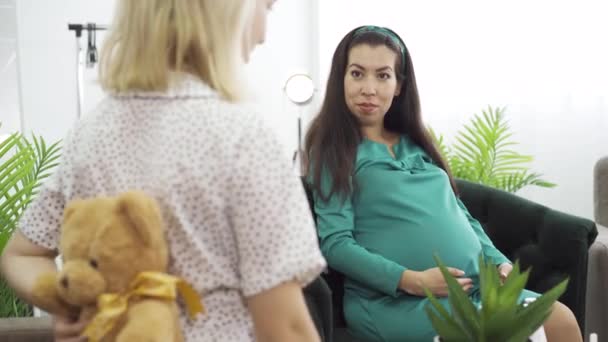 Dziewczyna w świetle kropkowana sukienka dając jej przyjaciel prezent dla jej nienarodzonego dziecka. Młoda brunetka przyszła Kobieta w niebiesko-zielonej sukni otrzymaniu misia na herpre-party urodzenia. — Wideo stockowe