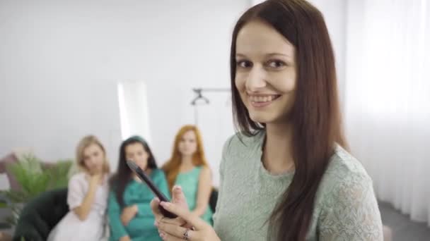 Joven chica sonriente en vestido azul-verde tomando fotos de tres amigos sentados en el entrenador. Compañía de mujeres que pasan tiempo con el expectante en la fiesta de pre-nacimiento. Centrado en el primer plano . — Vídeos de Stock