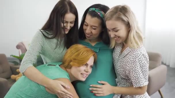 Cuatro mujeres caucásicas atractivas jóvenes abrazando y sonriendo. Chica pelirroja escuchando al niño en el vientre de sus amigos. Feliz compañía femenina teniendo fiesta de pre-nacimiento . — Vídeo de stock