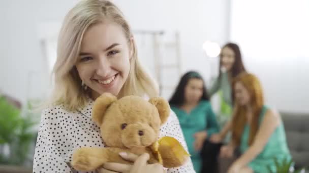 Primer plano de la joven rubia sosteniendo el osito de peluche y sonriendo. Compañía de sus amigos sentados en el fondo y charlando. Mujeres pasando tiempo juntas en interiores . — Vídeos de Stock