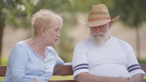Äldre kaukasiska par sitter på bänken i sommarparken och pratar. Positiv mogen man i halmhatt tillbringar tid med sin vackra fru utomhus. Livet efter pensioneringen. — Stockvideo
