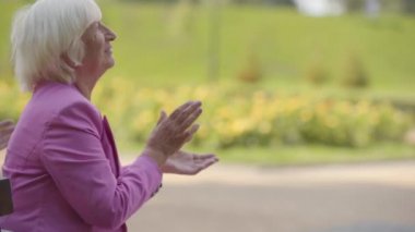 Sarışın, olgun bir kadının yaz parkında oturup ellerini çırpışının yan görüntüsü. Üst düzey bir erkek, onu dansa davet eden bir jestle ortaya çıkıyor. Emeklilikten sonraki aktif yaşam.