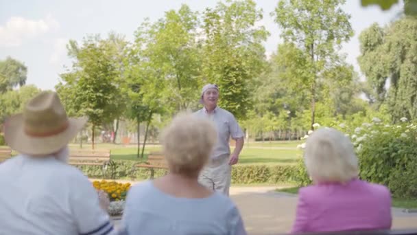 Glad kaukasisk senior man dansar framför sina vänner sitter vid bänken. Positiv gubbe som underhåller moget sällskap i sommarparken. Medelålders människor som njuter av bra väder. — Stockvideo