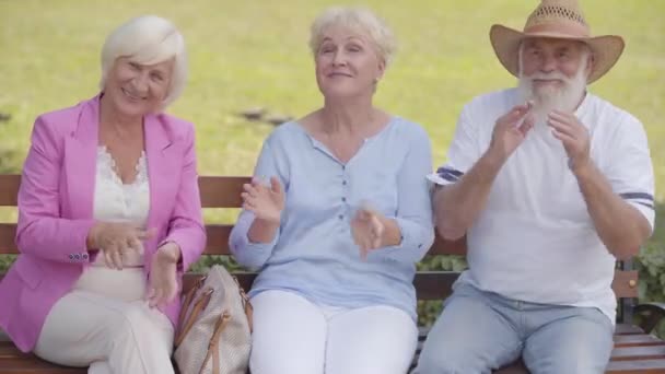 Three caucasian middle-aged people sitting at the bench and applauding. Elderly company having fun in the park after retirement. Two pleasant mature women and senior man in straw hat resting outdoors. — Stock Video