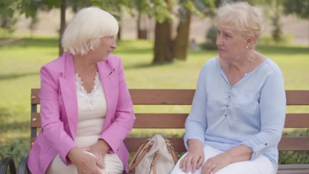 Pěkné dospělé bělošky sedící na lavičce v letním parku. Starší žena s blonďatými vlasy si stěžuje na bolest v koleni svému příteli. Starší ženy doporučující účinné léky proti bolesti. — Stock video