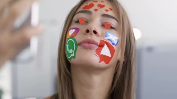 Κινούμενες κόκκινες καρδιές στα μάτια της νεαρής ελκυστικής γυναίκας με ζωγραφισμένες εικόνες κοινωνικών μέσων στο πρόσωπό της. Καυκάσιος κορίτσι στέλνει κινούμενα μηνύματα δεξιά από το πρόσωπό της σε τηλέφωνο και tablet. Εθισμός — Αρχείο Βίντεο