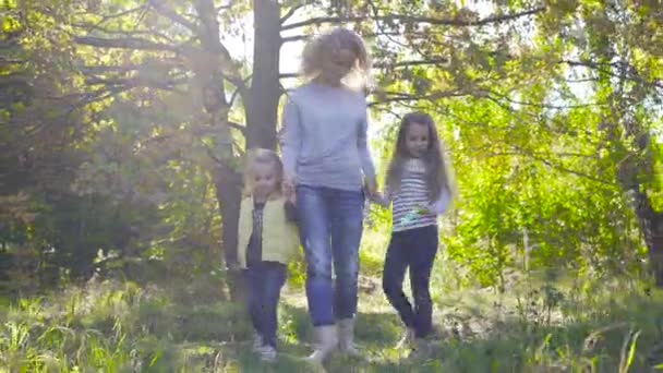 Boldog kaukázusi család sétál a napsugarakban az őszi parkban. Mosolygó, felnőtt, göndör nő, aki fogja a kislányai kezét. Nők alkalmi ruhák pihen a szabadban. — Stock videók