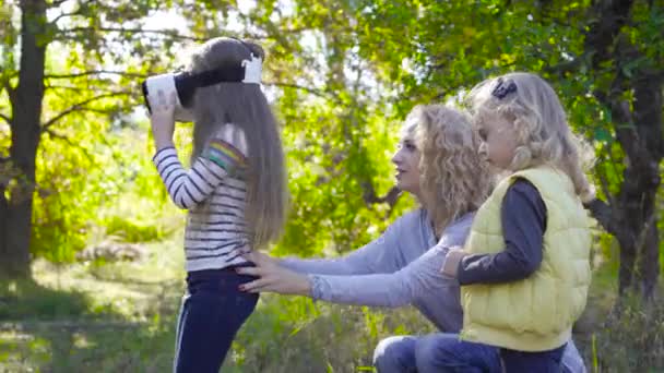 Vista lateral de una chica morena caucásica en gafas VR en el parque de otoño. Rubia sonriente madre caucásica apoyando a su hija y hablando con sus pequeños niños lindos . — Vídeos de Stock