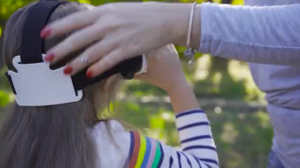 Gros plan de mains féminines caucasiennes mettant des lunettes VR sur la petite fille en chemisier rayé. Enfants jouant avec des casques de réalité virtuelle à l'extérieur . — Video