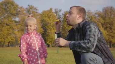 Beyaz adam küçük kızına sabun köpüğü üflemeyi öğretiyor. Babam, sonbahar parkında pembe noktalı ceket giymiş güzel sarışın kızı eğlendiriyor..