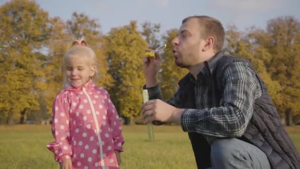 Kaukasischer Mann zeigt seiner kleinen Tochter, wie man Seifenblasen pustet. Vater unterhaltsam süße blonde Mädchen in rosa gepunktete Jacke im Herbst Park. — Stockvideo