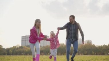 Küçük beyaz çocuk babasının ve ablasının elini tutuyor ve zıplıyor. Gündelik giysiler içindeki pozitif aile güz gününü dışarıda birlikte geçirir..