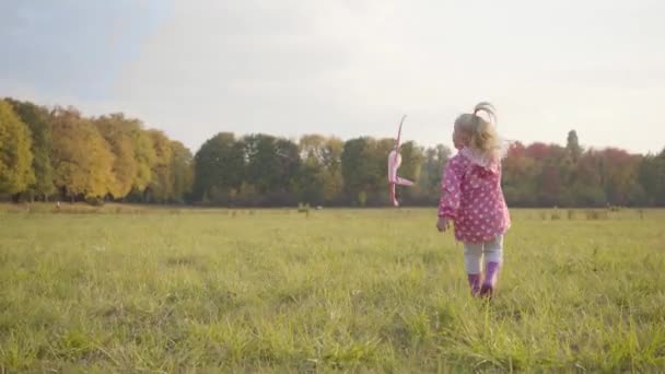 Kis szőke, fehér lány, pöttyös rózsaszín dzsekiben és gumicsizmában játszik egy játékgéppel a szabadban. Aranyos gyerek fut az őszi parkban este. — Stock videók