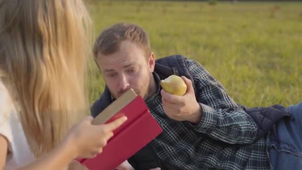 若い白人男性がリンゴを食べ、娘が本を読むのを待っている。父支出時間とともに若いですブロンドの女の子オンザ秋の牧草地. — ストック動画