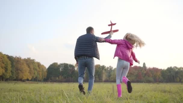 Visão traseira do homem caucasiano e menina loira jovem passeando e girando no prado do outono. Pai segurando avião de brinquedo rosa imitando sua mosca e sua filha correndo ao lado dele e rindo . — Vídeo de Stock