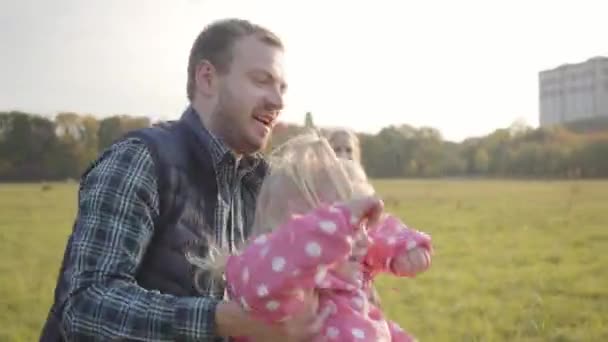 Mladý běloch, který na podzim večer venku točí roztomilé dítě rukama. Otec baví své dvě blond dcery oblečené v růžových šatech. — Stock video