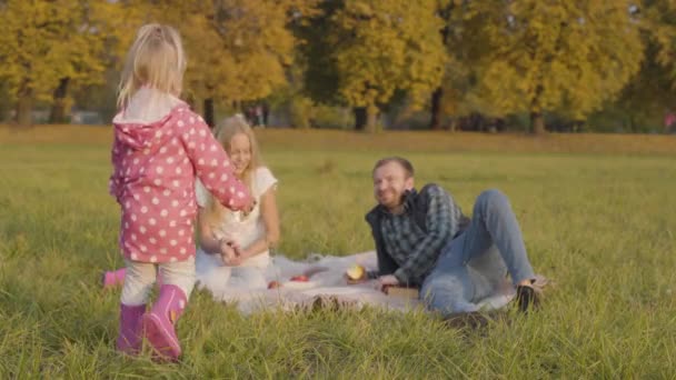 Kleines blondes Kind in pinkfarbener Kleidung, das zu der älteren Schwester und dem Vater geht, die auf der Herbstwiese sitzen und Äpfel essen. glückliche kaukasische Familie beim Picknick im Freien. — Stockvideo