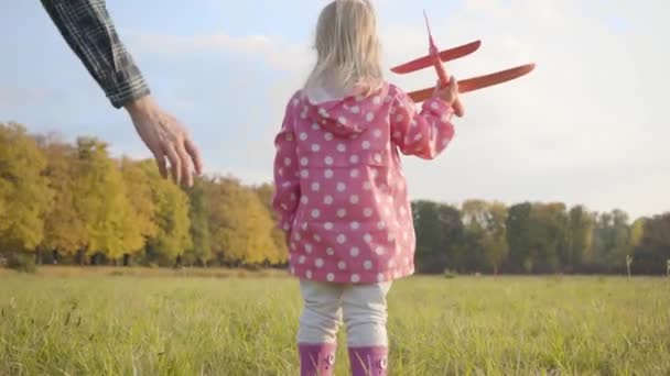 Jeune homme caucasien aidant son adorable petite fille à jouer avec un avion jouet rose. Jolie fille blonde souriante en veste pointillée et bottes en caoutchouc courant avec son père dans la soirée d'automne . — Video