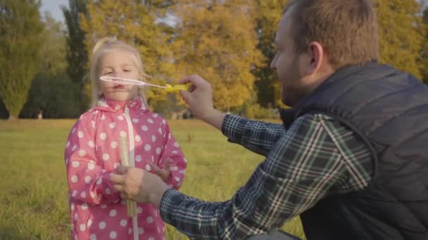 Позитивний кавказький чоловік грає зі своєю маленькою донькою в осінньому парку. Симпатична блондинка одягнена в рожевий пунктирний піджак, що дме мильні бульбашки. Дитина проводить час з батьком на відкритому повітрі .. — стокове відео