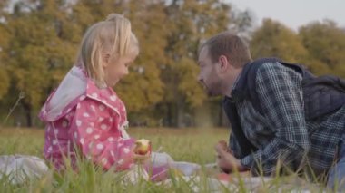 Noktalı pembe ceketli küçük kız ve çayırda oturup elma yiyen yetişkin beyaz adam. Baba güz gününü sevgili kızıyla dışarıda geçiriyor..