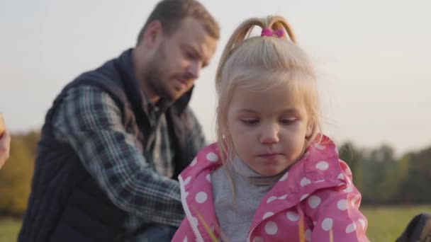 Ritratto ravvicinato di una bambina carina con i capelli biondi e gli occhi marroni che guardano la macchina fotografica. Bella bambina che ascolta suo padre seduto sul prato autunnale e parla . — Video Stock