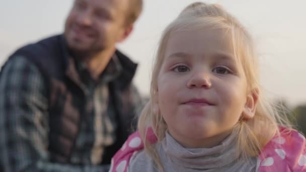 Detailní záběr malé holčičky s blonďatými vlasy a hnědýma očima, dívající se do kamery. Hezké dítě sedí se svým otcem na podzimní louce. Zaměřeno na popředí. — Stock video