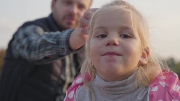 Närbild av en liten söt flicka med blont hår och bruna ögon tittar på kameran. Ett vackert barn som sitter med sin far på höstängen. Fokuserat på förgrunden. — Stockvideo