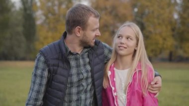 Gülümseyen beyaz bir adam ön plandaki genç sarışın kıza yaklaşıp ona sarılıyor. Baba, sonbahar parkında pembe ceketli ergen kızıyla bir şeyler yapıyor..